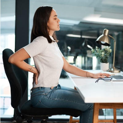 Na zdjęciu widzimy kobietę siedzącą przy biurku w nowoczesnym biurze, wyraźnie odczuwającą dyskomfort w dolnej części pleców – sygnalizuje to ręka oparta na krzyżu i lekko pochylona sylwetka. Kobieta ma na sobie jasną koszulę i dżinsy, siedzi na ergonomicznym czarnym krześle, przy biurku z laptopem. Obok komputera widać okulary, lampkę i małą doniczkę z rośliną. Tło stanowią duże okna oraz nowoczesne wyposażenie biura.