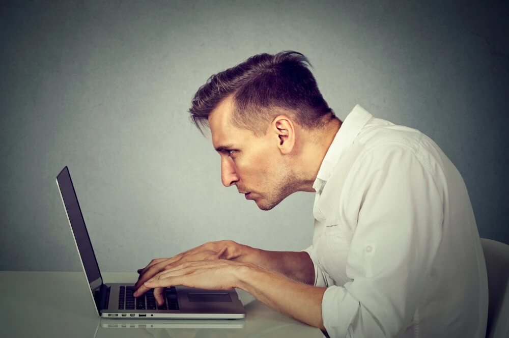 Zdjęcie przedstawia mężczyznę, który siedzi przy laptopie w bardzo złej pozycji. Głowa mężczyzny jest mocno wysunięta do przodu, a plecy są zgarbione.