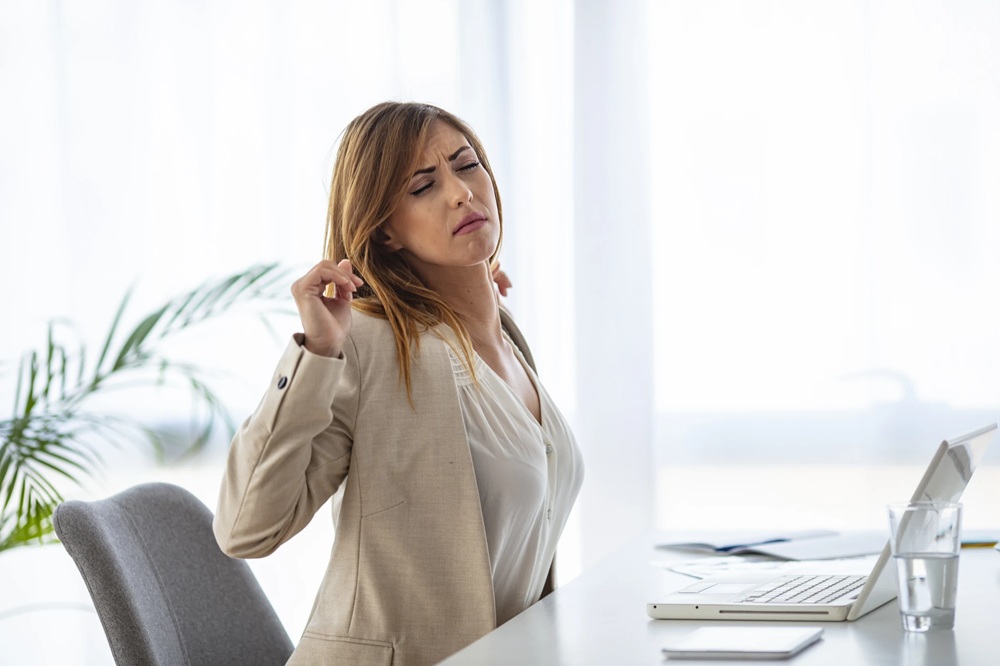 Na zdjęciu widzimy kobietę ubraną w strój biznesowy, siedzącą przy biurku w jasnym, nowoczesnym pomieszczeniu. Przed nią znajduje się laptop, szklanka wody oraz dokumenty, co sugeruje pracę biurową lub zdalną. Kobieta reaguje na dyskomfort – prawdopodobnie w okolicach karku lub górnej części pleców. W tle widać roślinę doniczkową.