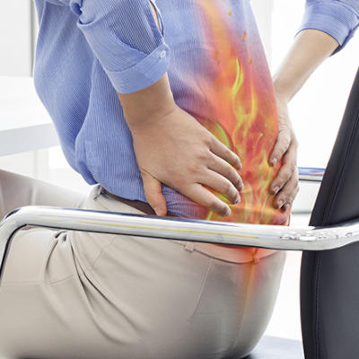 Na zdjęciu widzimy osobę siedzącą na krześle, trzymającą się za dolną część pleców, co wyraźnie wskazuje na silny ból krzyża. Obszar lędźwiowy został dodatkowo zaznaczony graficzną ilustracją płomieni, symbolizującą intensywne uczucie pieczenia lub dyskomfortu. Osoba ubrana jest w neutralne kolory, a tło pozostaje rozmyte, by skupić uwagę na problemie bólu pleców.